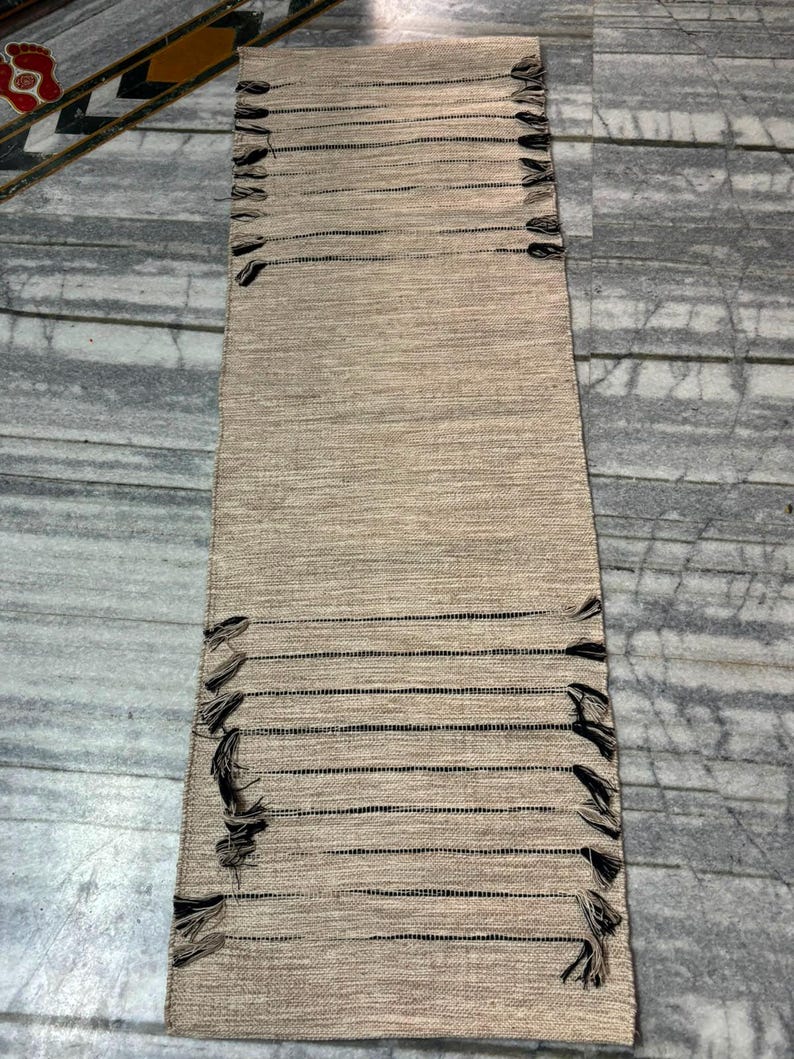 May include: A rectangular woven table runner in a natural beige hue, featuring horizontal dark brown thread accents and decorative fringe detailing along the side edges. The runner has a textured, fibrous appearance with a consistent weave pattern.