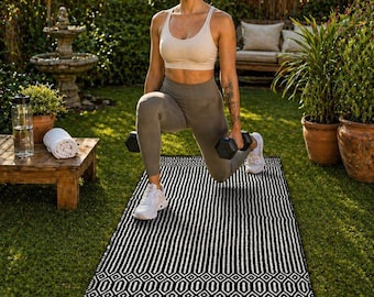 May include: A black and white patterned yoga mat is laid out on a green lawn. The mat has a geometric design at the bottom and vertical stripes in the center. A person is doing lunges on the mat, holding dumbbells.