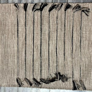 May include: A rectangular woven rug in a neutral beige tone featuring a series of vertical black stripes. The rug is accented with decorative tassels at both the top and bottom ends of each black stripe, creating a textured and linear pattern.