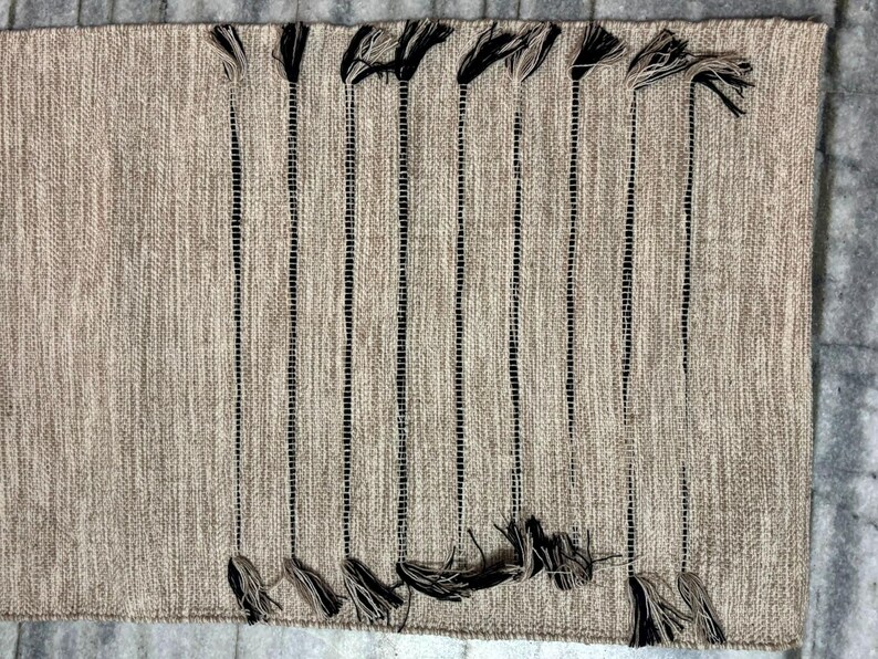 May include: A rectangular woven rug in a neutral beige tone featuring a series of vertical black stripes. The rug is accented with decorative tassels at both the top and bottom ends of each black stripe, creating a textured and linear pattern.