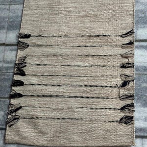 May include: A rectangular table runner featuring a woven beige fabric with a textured surface. The runner includes horizontal black thread accents and decorative fringe tassels in black and beige along both of the long side edges.