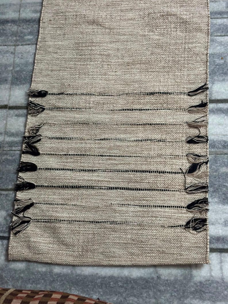 May include: A rectangular table runner featuring a woven beige fabric with a textured surface. The runner includes horizontal black thread accents and decorative fringe tassels in black and beige along both of the long side edges.