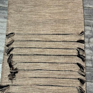 May include: A rectangular table runner featuring a woven tan texture with horizontal black stripes. The design includes decorative fringe tassels attached to the edges of the black stripes, creating a textured appearance across the surface.