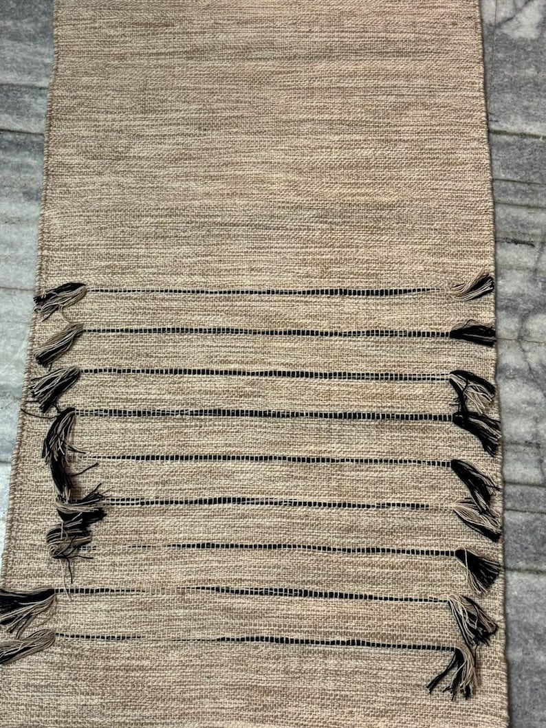 May include: A rectangular table runner featuring a woven tan texture with horizontal black stripes. The design includes decorative fringe tassels attached to the edges of the black stripes, creating a textured appearance across the surface.