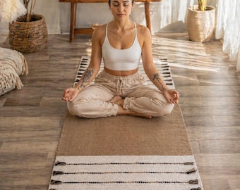 May include: A woman meditating on a rectangular yoga mat. The mat is primarily brown with a cream-colored section featuring horizontal black and brown stripes. The mat has tassels at the end. The woman is wearing a white top and beige pants.