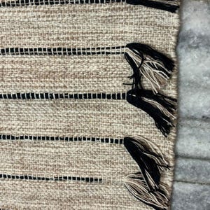 May include: A woven textile rug featuring a textured beige base with horizontal black stripes. The edge of the rug is finished with decorative tassels that combine black and beige fibers, creating a distinct contrast against the woven fabric.