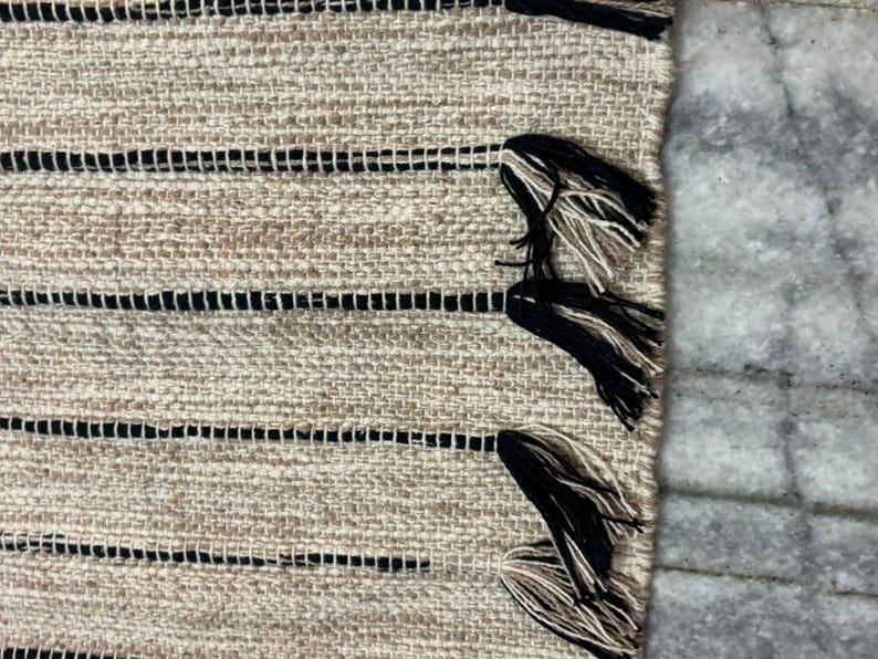 May include: A woven textile rug featuring a textured beige base with horizontal black stripes. The edge of the rug is finished with decorative tassels that combine black and beige fibers, creating a distinct contrast against the woven fabric.