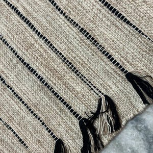 May include: A woven rug featuring a textured beige and tan base with prominent vertical black stripes. The edge of the rug is finished with decorative tassels composed of alternating black and beige yarn strands, creating a distinct pattern.