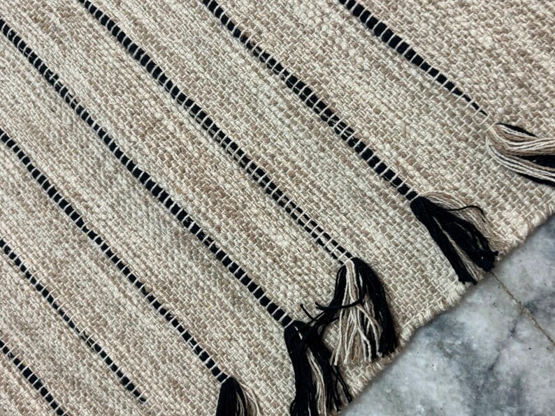 May include: A woven rug featuring a textured beige and tan base with prominent vertical black stripes. The edge of the rug is finished with decorative tassels composed of alternating black and beige yarn strands, creating a distinct pattern.