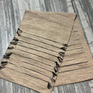 May include: A rectangular table runner in a light tan, textured fabric featuring horizontal black stripes. The edges of the runner are finished with small, dark brown tassels attached at regular intervals along both of the longer sides.