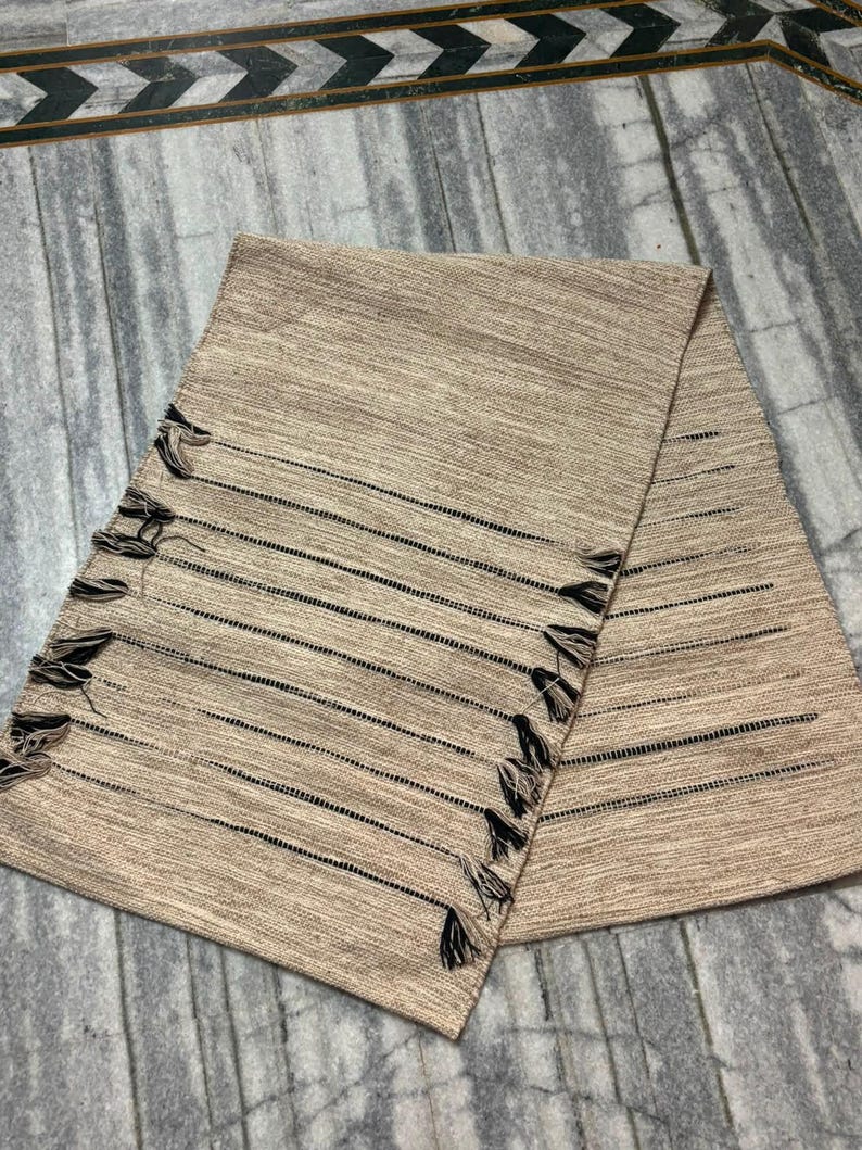 May include: A rectangular table runner in a light tan, textured fabric featuring horizontal black stripes. The edges of the runner are finished with small, dark brown tassels attached at regular intervals along both of the longer sides.
