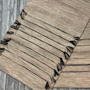 May include: A rectangular woven table runner in a light tan color featuring a series of thin, horizontal black stripes. The edges of the runner are decorated with small, dark-colored tassels that are evenly spaced along the length of the fabric.