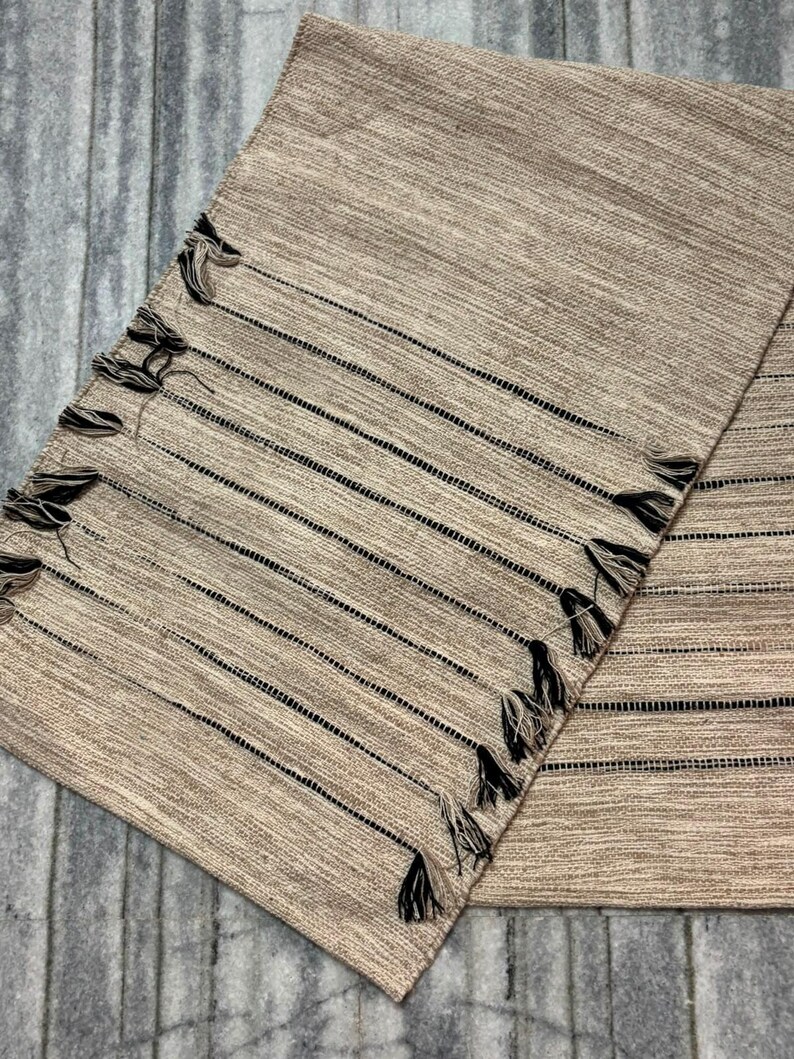 May include: A rectangular woven table runner in a light tan color featuring a series of thin, horizontal black stripes. The edges of the runner are decorated with small, dark-colored tassels that are evenly spaced along the length of the fabric.