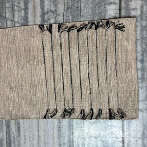 May include: A rectangular woven table runner in a light beige, textured fabric. The runner features a series of vertical, thin black stripes running across the surface, each ending in a small, dark tassel at both the top and bottom edges.