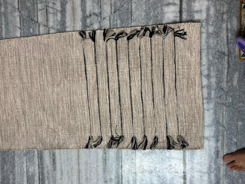May include: A rectangular woven table runner in a light beige, textured fabric. The runner features a series of vertical, thin black stripes running across the surface, each ending in a small, dark tassel at both the top and bottom edges.