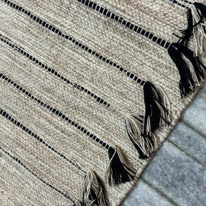 May include: A woven area rug featuring a textured tan and beige surface with thin, vertical black stripes. The edge of the rug is finished with a series of decorative tassels, each composed of a blend of black and tan fibers hanging from the stripes.