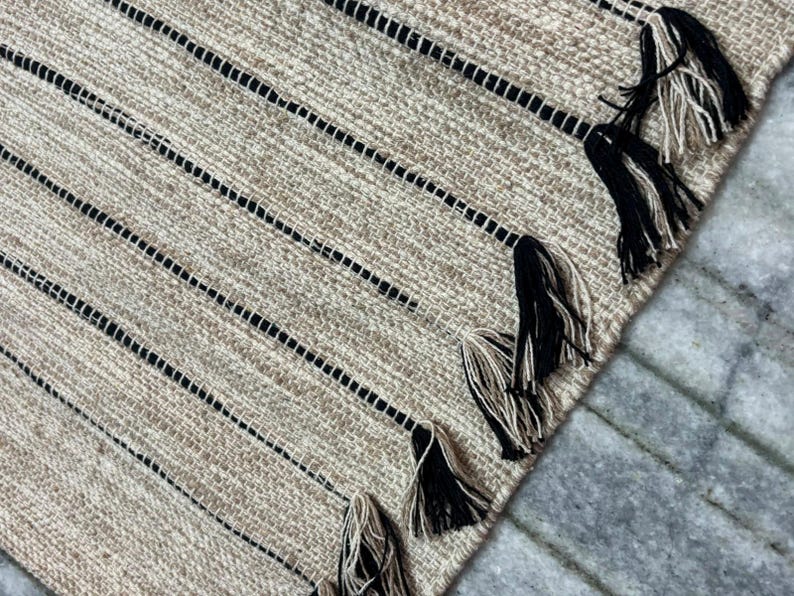 May include: A woven area rug featuring a textured tan and beige surface with thin, vertical black stripes. The edge of the rug is finished with a series of decorative tassels, each composed of a blend of black and tan fibers hanging from the stripes.