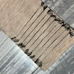 May include: A rectangular table runner in a light tan, woven fabric featuring a series of thin, dark brown vertical stripes. The edges of the runner are finished with a decorative fringe made of dark brown yarn tassels along the short sides.