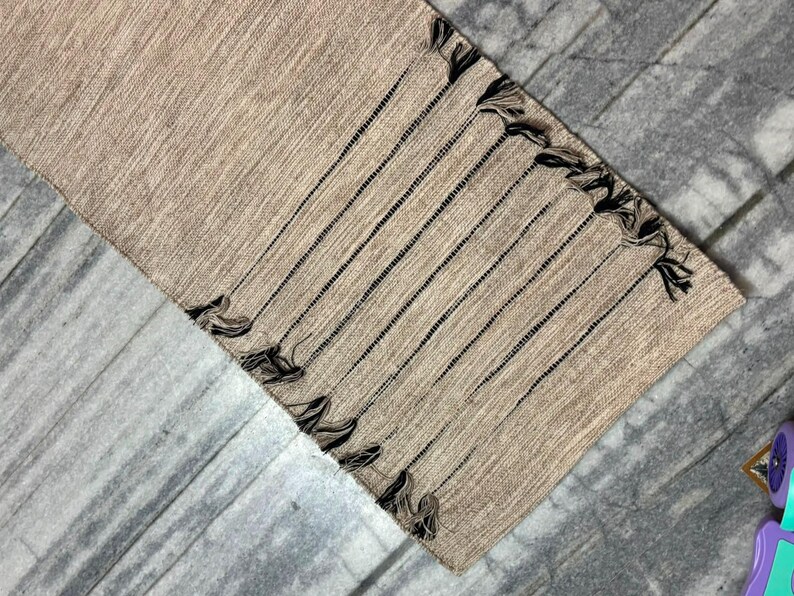 May include: A rectangular table runner in a light tan, woven fabric featuring a series of thin, dark brown vertical stripes. The edges of the runner are finished with a decorative fringe made of dark brown yarn tassels along the short sides.
