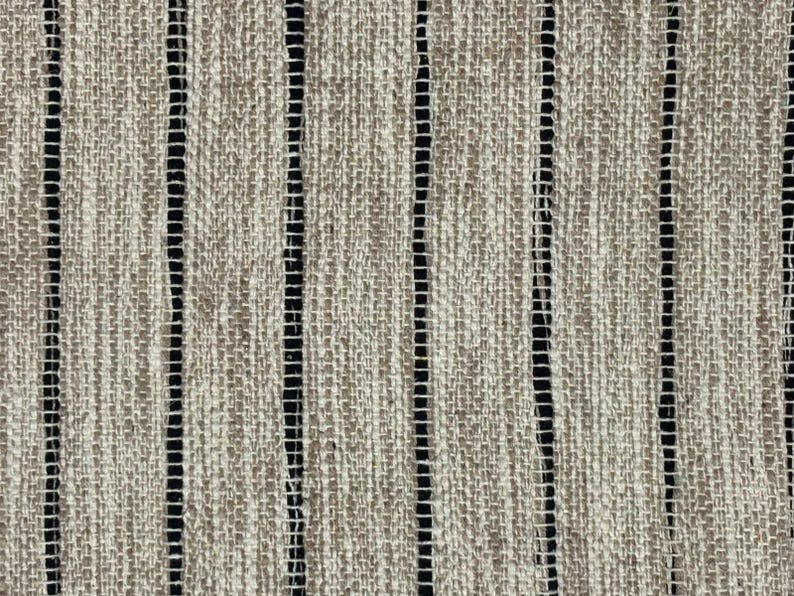 May include: A woven fabric featuring a textured, light brown and beige surface with a prominent vertical striped pattern. The design consists of alternating wide, coarse-textured tan bands and thin, dark black vertical lines spaced evenly throughout.