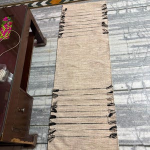 May include: A rectangular woven runner rug featuring a natural beige textured surface with horizontal black thread accents. The edges of the rug are finished with frayed black fringe details along both sides, creating a linear pattern.