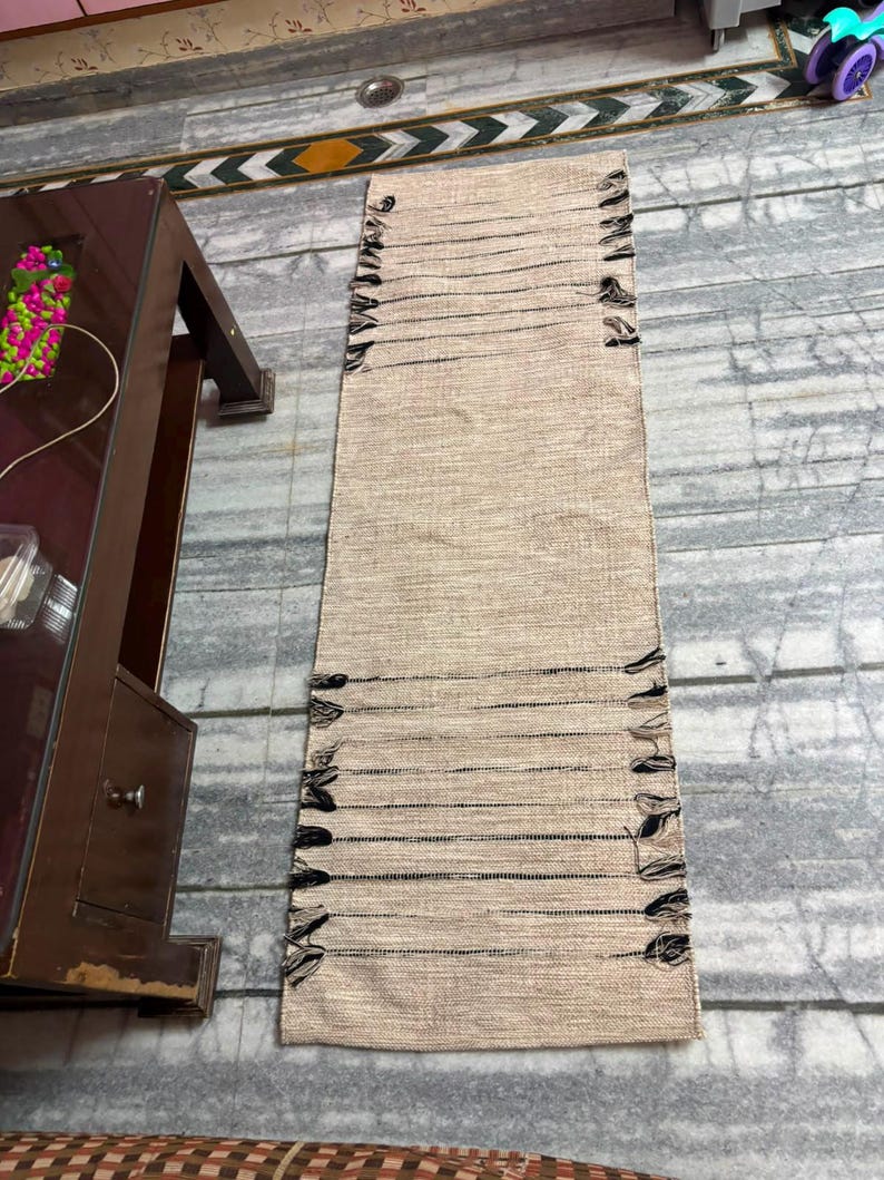 May include: A rectangular woven runner rug featuring a natural beige textured surface with horizontal black thread accents. The edges of the rug are finished with frayed black fringe details along both sides, creating a linear pattern.