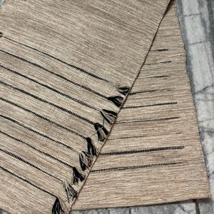 May include: A rectangular beige table runner featuring a woven texture with thin horizontal black stripes. The fabric includes openwork detailing along the stripes and a series of small, dark-colored tassels attached along one of the diagonal edges.