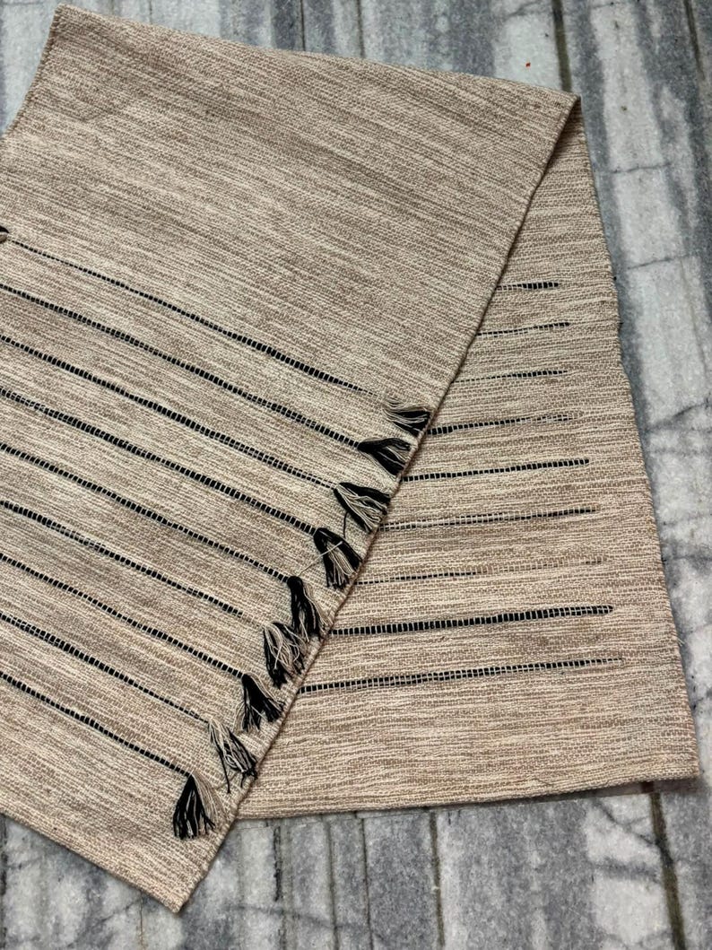 May include: A rectangular beige table runner featuring a woven texture with thin horizontal black stripes. The fabric includes openwork detailing along the stripes and a series of small, dark-colored tassels attached along one of the diagonal edges.