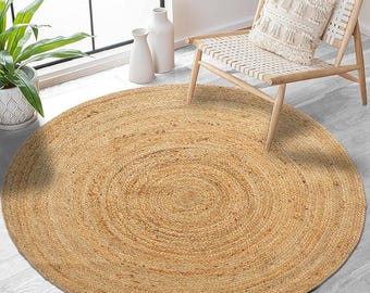 Puede incluir: Una alfombra redonda de yute en tonos naturales con un diseño en espiral. La alfombra está colocada sobre un suelo de madera gris claro, con una silla de madera y una ventana al fondo. Se aprecia la textura tejida de la alfombra.