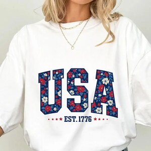 May include: A white short-sleeved t-shirt featuring the text USA in large block letters filled with a red, white, and blue floral pattern. Below the letters, the text EST. 1776 is printed in blue, flanked by four small red stars on each side.
