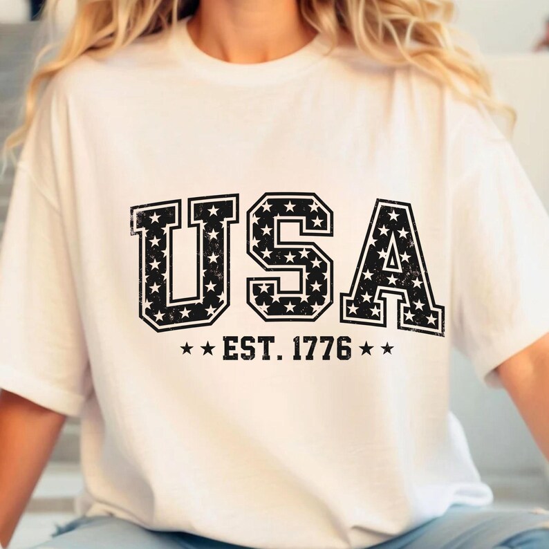 May include: A white short-sleeved t-shirt featuring the black text USA in a block font with a distressed star pattern inside the letters. Below the text, the words EST. 1776 are printed in black, flanked by two small black stars on each side.