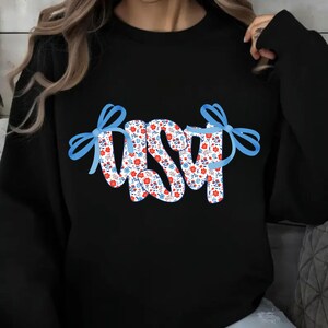 May include: A black long-sleeve sweatshirt featuring the letters USA in a white font filled with a repeating pattern of small red and blue flowers. The letters are accented on both sides by large, light blue decorative ribbon bows.