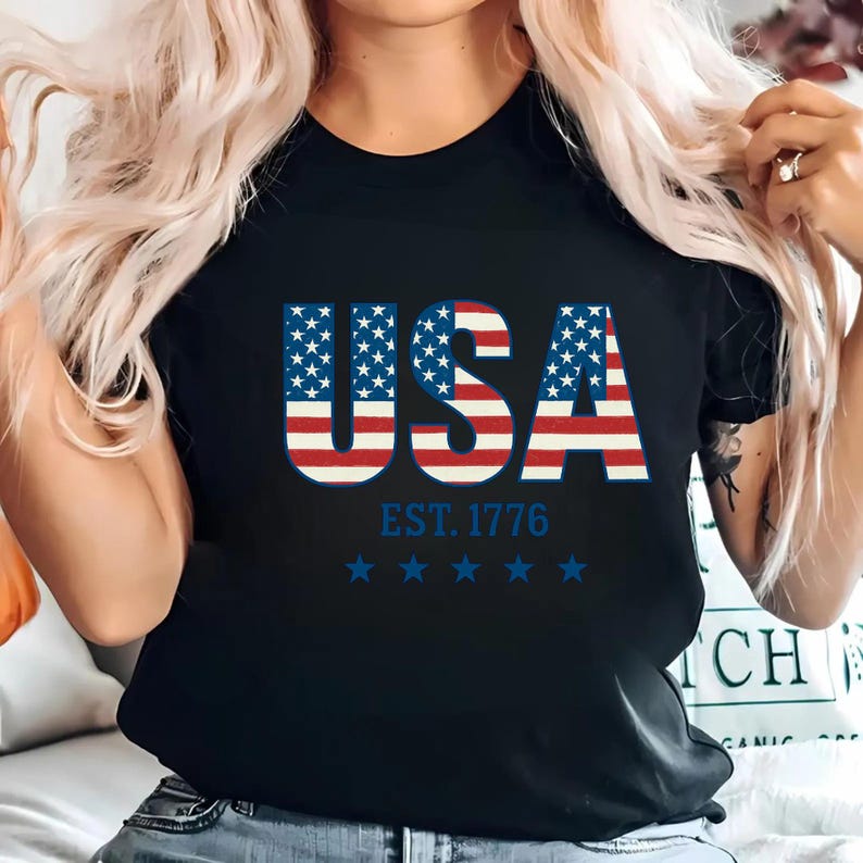 May include: A black short-sleeved t-shirt featuring the text USA in a red, white, and blue American flag pattern. Below the text, the shirt displays the blue text EST. 1776 followed by a horizontal row of five blue stars centered on the chest.