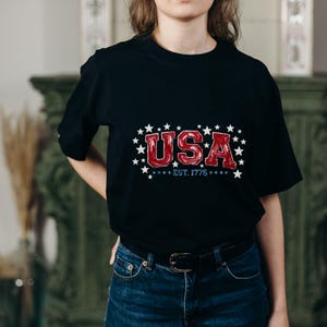 May include: A black short-sleeved t-shirt featuring a graphic print with the red text USA, the blue text EST. 1776, and a surrounding pattern of white stars. The shirt is paired with dark blue denim jeans secured by a black leather belt.