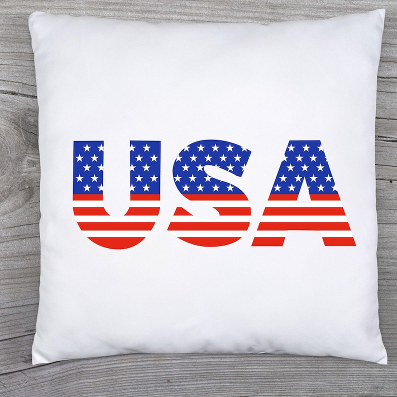 May include: A square white throw pillow featuring the letters USA printed in the center. The letters are filled with a pattern of white stars on a blue background on the top half and horizontal red and white stripes on the bottom half.