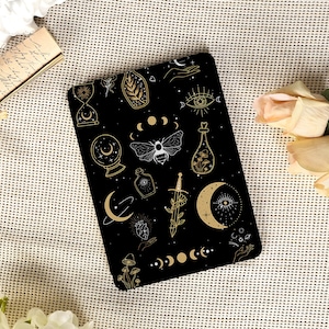 Op de afbeelding: Een zwart notitieboek met gouden en witte hemelse illustraties. Het ontwerp bevat manen, sterren, een kristallen bol, een mot en andere mystieke symbolen. Het notitieboek staat tegen een neutrale, getextureerde achtergrond.