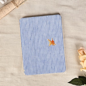 Può includere: Un taccuino rettangolare a righe blu e bianche con un'illustrazione di un pesce rosso arancione. Il taccuino è appoggiato su una superficie beige strutturata, con una pergamena arrotolata e una rosa.