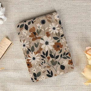May include: A rectangular notebook with a floral pattern featuring white daisies, brown accents, and green leaves on a beige background. The notebook is surrounded by a neutral-toned surface.