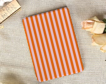May include: A rectangular notebook with a vertical striped pattern in orange and light pink. The notebook is on a textured, off-white surface, with floral accents and a vintage-style fan nearby.