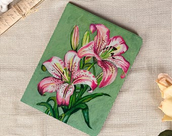 May include: A green journal with a hand-painted floral design featuring pink and white lilies. The lilies have dark pink spots and green leaves. The journal is on a white textured surface.