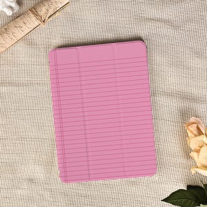 May include: A pink tablet case with a lined paper design. The case is rectangular and has a smooth surface. The background is a textured, off-white fabric. A rolled-up scroll and a rose are also in the image.