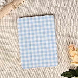 Op de afbeelding: Een lichtblauw en wit geruit patroon tablethoesje. De hoes is rechthoekig en heeft afgeronde hoeken. De achtergrond is een beige geweven stof. Een opgerolde rol en een roos staan ook in de afbeelding.