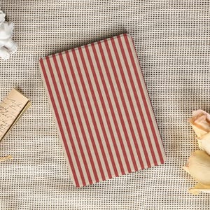 May include: A rectangular notebook featuring a red and cream striped design. The notebook rests on a textured, off-white surface. Decorative elements, including a small angel statue and dried flowers, are in the background.