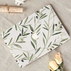 May include: A laptop cover featuring a watercolor design of olive branches with green leaves and black and green olives. The cover has a white background and a cutout for the Apple logo.