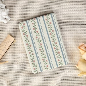 Puede incluir: Un cuaderno rectangular con un estampado floral y de rayas. El diseño presenta rayas verticales de color azul claro con un borde ondulado, alternando con un fondo crema adornado con flores rosas y hojas verdes.