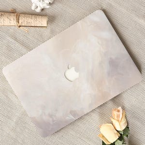 Puede incluir: Un ordenador portátil con una cubierta de diseño abstracto de color claro. La cubierta tiene un patrón similar al mármol en tonos de blanco, beige y gris. El logotipo de Apple es visible en la parte superior del portátil.