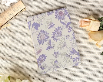 Puede incluir: Un cuaderno rectangular con un diseño floral. La cubierta presenta un fondo gris claro con ilustraciones de flores moradas y blancas. El cuaderno está rodeado de flores y una carta de estilo vintage.