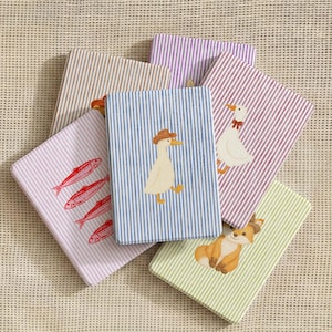 May include: A collection of six rectangular notebooks with striped covers in various colors including blue, purple, red, beige, and green. Each notebook features a small illustration, such as a duck wearing a hat, a goose, a fox, and red fish.