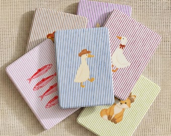May include: A collection of six rectangular notebooks with striped covers in various colors including blue, purple, red, beige, and green. Each notebook features a small illustration, such as a duck wearing a hat, a goose, a fox, and red fish.