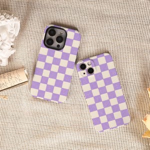 May include: Two phone cases featuring a purple and white chequered pattern. The cases are designed for a smartphone, with camera and button cutouts. The cases are displayed on a textured surface.